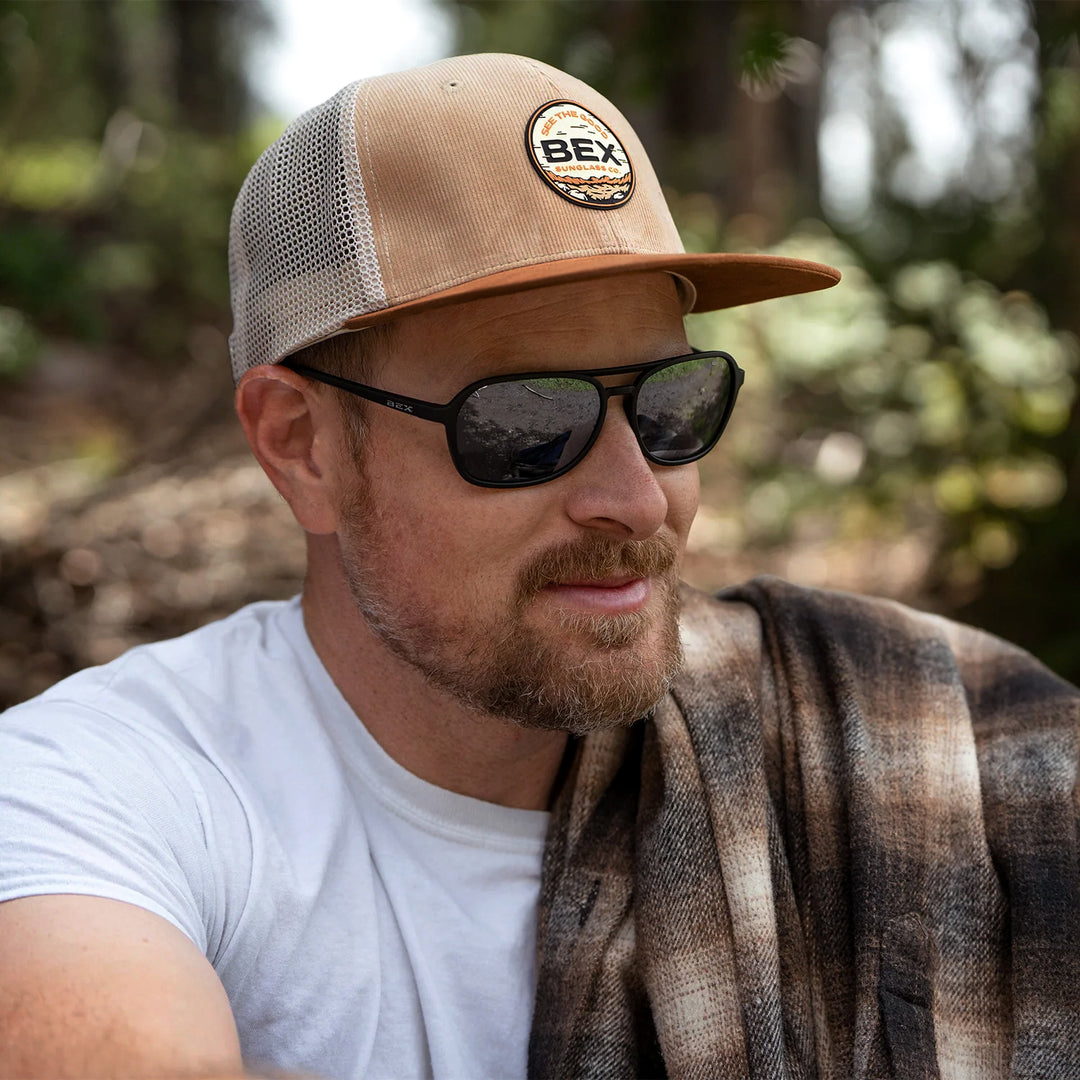 Man wearing a tan flat bill hat with a BEX front patch outdoors #color_tan-brown